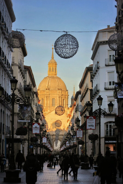 A vibrant street-level view looking down a busy pedestrian avenue lined with tall, classic white buildings featuring balconies. In the centre distance, a magnificent basilica dome and spire are bathed in warm, golden sunset light, contrasting with the shadowed street below. Large, wire-frame spherical holiday decorations hang suspended across the sky between the buildings. The street is crowded with the silhouettes of people walking, and ornate black lampposts with banners line the walkway.