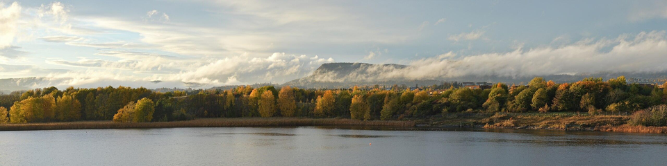 A panoramic photo of a bay with a forest with it's leaves turning yellow on the far shore. There are low mountains in the distance with clouds drifting over them.
