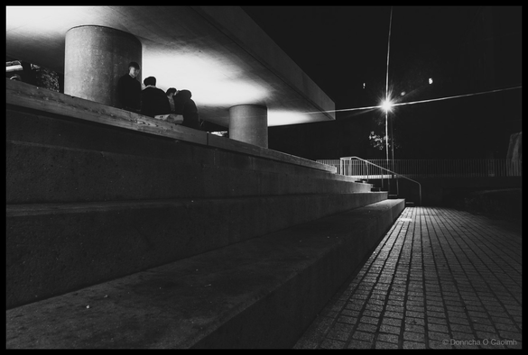 Black and white night-time photograph of several figures sitting on a raised concrete platform near cylindrical concrete pillars and concrete roof, dramatic fluorescent lighting creating stark contrast against dark sky, modern metal railing visible on right, brick paved ground in foreground, taken at Bishop Lucey Park, Cork.
