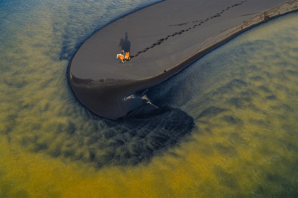 Aerial view of a person riding a horse on a black sandbank surrounded by rippling blue and green water.