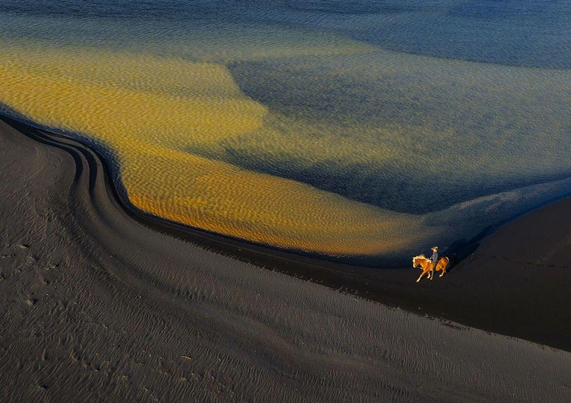A solitary horse and rider walk along a black sand beach, with yellowish water flowing gently nearby, creating a serene landscape.