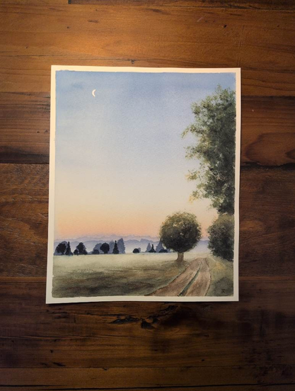 This is a watercolor landscape painting in portrait orientation on Saunders Waterford Coldpress watercolor paper in 8x10. This is a landscape of a sky at sunset with a little crescent moon in the top left corner of the medium value warm blue sky.  This color fades down into a lighter blue, then at the horizon line is a peachy pink and yellow swatch of sky. The background are layers of misty blueish tree lines that get darker as they move forward. Then there is a stretch of lawn with a little road on the ride side that disappears at the horizon line. The top of the lawn is a pale green giving a misty appearance which comes more into focus as it moves to the foreground. At the mid ground there's a tree on the left side of the road. On the right side there is some green foliage that takes up almost the entire right side.