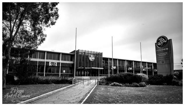Black and white photograph of the long, two story administrative building for the former Holden (GMH) Manufacturing Operations, featuring a large circular Holden lion emblem above the entrance and a tall sign to the right displaying the Holden logo. A pathway leads up to the building entrance, flanked by landscaping.