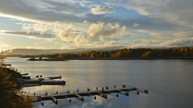 A photo of a bay with forests with yellow leaves on the far shore. The bay has several docks in it. The sky is blue with many clouds in it and the sun is settings.