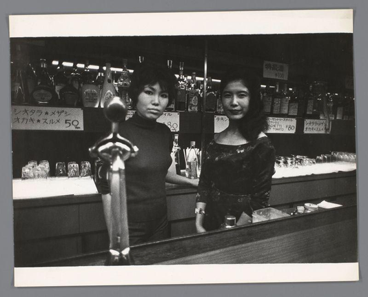 The image depicts two women standing behind a bar counter. The woman on the left is wearing a black, turtleneck dress and has short hair that appears to be dark in color. She stands next to another individual who also seems to have dark hair but wears what looks like an ornate top with patterns or embellishments.

Behind them are shelves stocked with various bottles of liquor, some labeled as Japanese sake, indicating a bar setting likely located in Japan due to the writing on the labels and signage which is written in Japanese characters. On the counter, there's pricing for drinks such as "かめお酒" priced at 100 yen or "サStra**マ**ン," along with other items like "ガチetti ¥80." These prices suggest a casual dining environment.

The woman on the left appears to be holding onto what looks like a beer tap handle, suggesting that she might be an attendant or bartender. The bar has several glasses lined up neatly in front of them which are presumably for serving drinks. There's also another person visible through a mirror reflection at their side, but they're not fully shown.

The image is presented as a black and white photograph with no coloration except for the grayscale tones typical to such imagery. It captures an authentic moment likely from everyday life in Japan during the early part of 20th century based on its style and content.