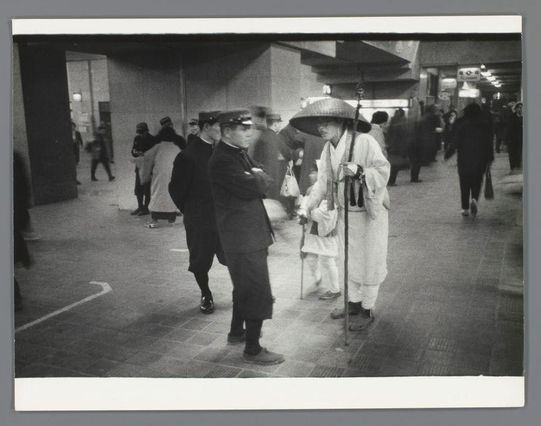 The image shows a scene inside what appears to be an urban transportation hub, likely a train station. In the foreground, two individuals are engaged in conversation; one is dressed in traditional Japanese attire and holding a bamboo walking stick or umbrella, while the other wears Western-style formal wear with a peaked cap indicative of staff uniforms from that era.

The background reveals several more people moving about their business, some blurred due to motion, suggesting movement within the space. The architecture has simple lines, large pillars supporting an overhead structure, and signage indicating directions for various exits or platforms is visible in one corner. It's likely a public place with high foot traffic during this period.

The monochrome quality of the photograph suggests it was taken at least several decades ago when color photography had not yet become widespread. The photo captures a moment that highlights cultural diversity within Japan, blending traditional and modern elements as people interact or pass by in their daily routines.