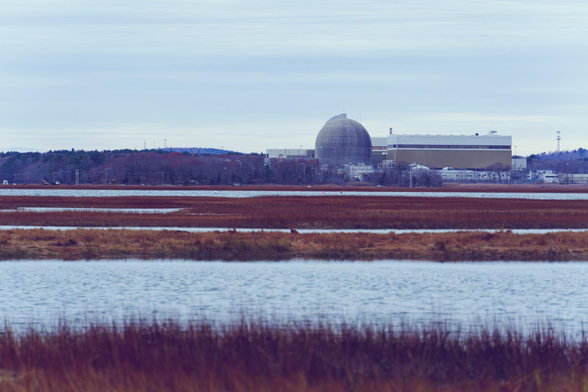 The horizon is near the center of this image and a nuclear power plant with a dome and a big turbine house sticks out from it,  above it some striated clouds and below a beautiful salt marsh