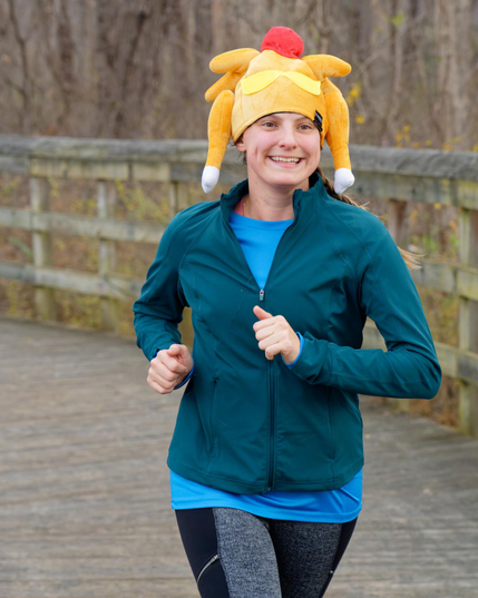 Woman in a blueish green jacket has a brighter blue shirt and black and grey pants and a big yellow, white and red turkey hat with a wood slat bridge in back