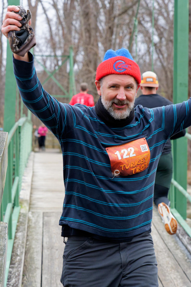 Man with red and blue hat and a dark blue shirt with pairs of horizontal light blue stripes and bib number 122 holds his hands up high at the close end of a pedestrial suspension bridge