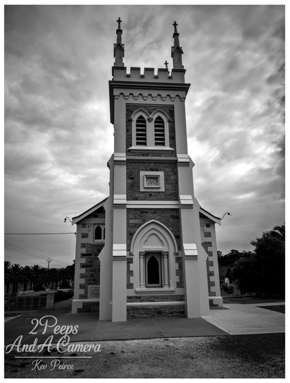 A black and white, low angle photograph of the historic Manaranga Church, featuring a prominent central tower with Gothic style pointed arch windows and battlements at the top, capped by two small spires.

The church facade is built from contrasting materials, possibly stone and render. The sky is heavily clouded, adding a dramatic, moody atmosphere.