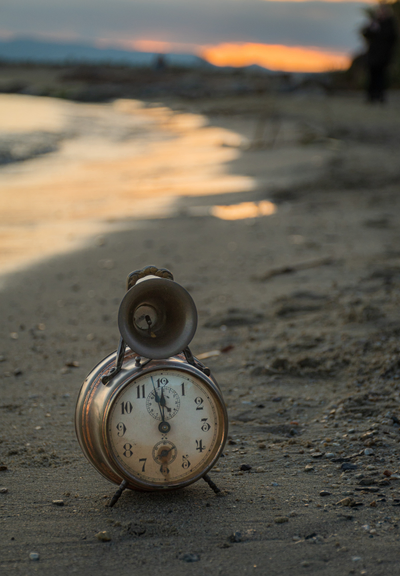 Das Bild zeigt einen alten, leicht angelaufenen Wecker, der am Strand im Sand steht. Der Hintergrund ist ein Sonnenuntergang über dem Meer. Die Farben des Sonnenuntergangs sind in warmen Tönen gehalten, was dem Bild eine ruhige und besinnliche Atmosphäre verleiht. Die Uhr zeigt kurz vor 12 Uhr an.