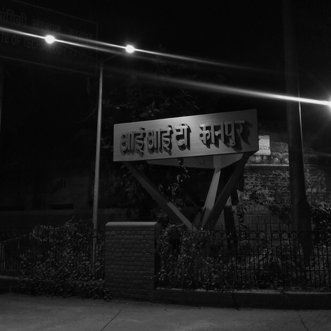 A night scene with a sign reading "IIT Kanpur" in Hindi, illuminated by streetlights. (black and white)