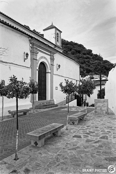 A narrow square lined with small trees and stone benches and Iglesia de Santo Domingo de Guzmán, featuring an arched doorway. A hill rises in the background, creating a serene, picturesque atmosphere in black and white.