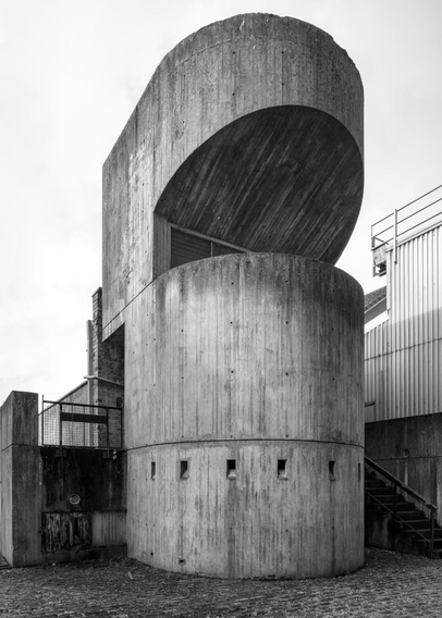 A huge concrete ventilation pillar (? it's not explained) rising from the ground by stairs that lead to the upper level of the building.

From the Guardian caption:

' Thomas C Keay Ltd (former offices), Dundee, designed by Ronald Cuddon with Tandy, Halford and Mills, built 1970

Many of the buildings constructed in the socially motivated period of post-war architecture have now been repurposed, pulled down or left to slowly decay. But others still serve their community. These stadiums and homes, leisure centres and fire stations, churches and libraries, were built for a people and nation in flux, with the vision of a new era of opportunity. '