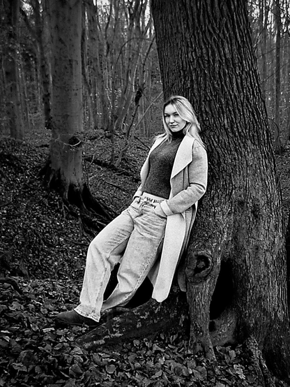 A young woman leans relaxed against a tree trunk in a forest. She wears a long coat, a sweater, and wide trousers. Her hands are in her pockets, and she stands on leaves covering the forest floor. More leafless trees are visible in the background.