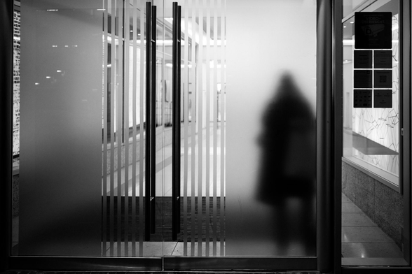 The image is a black and white photograph depicting an entranceway. The composition is primarily defined by glass doors, partially frosted, and a visible interior space.

The main subject is the silhouette of a person standing behind the frosted glass. The silhouette is dark and undefined, adding an element of mystery or anonymity. The glass door has vertical frosted stripes, creating a repeating pattern that leads the eye into the interior.