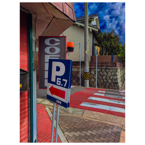 Description for the visually impaired: A color photo of a street corner featuring a blue and white parking sign reading “P 6.7” with a red arrow pointing left. Behind it, a vertical “COFFEE” sign hangs beside a red brick wall. A crosswalk, utility pole, and residential houses fill the background under a vivid blue sky.
