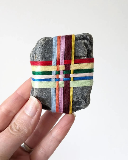Textile art, consisting of a small bit of weaving wrapped around a small stone. In this photo, the artist is holding up a small, squarish rock, which has colorful threads woven around it, similar to how one would wrap a box with a ribbon.