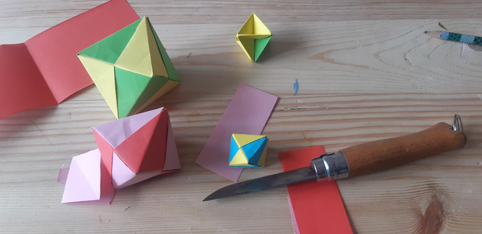 Photo d'origamis sur une table en bois. Polyèdres à 8 faces, en damier de couleurs. Mon fide opinel est présent por le découpage. 