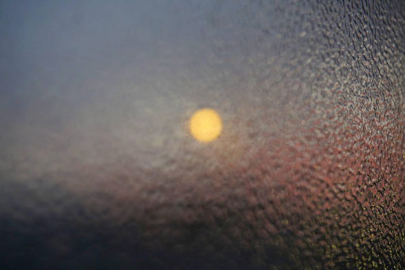 The photograph shows a hazy light that appears as a bright, blurred spot. The background is rendered in delicate shades of purple and blue, suggesting that this may be a scene at sunrise or sunset. The blurring effect gives the impression that the image could have been taken through wet or frozen glass, giving it a soft, dreamlike quality.