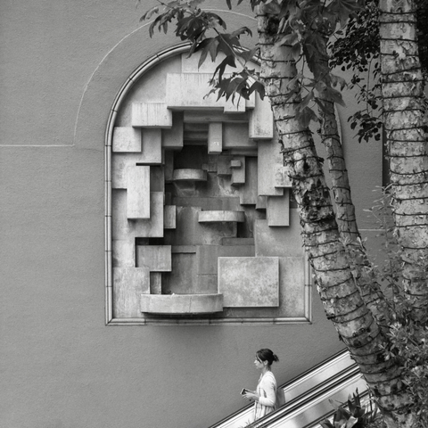 A black-and-white film photo. A woman is descending an escalator in front of a large art installation on the side of the building in downtown Los Angeles, California. Saturday, May 17, 2025.