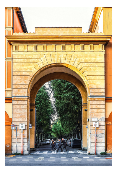 The photo is of city road framed by an archway, with pedestrians crossing it. The arch is painted in shades of yellow and orange, which frames the street. It is set against buildings with a similar colour palette.
Inside, through the arch, a street lined with tall, green trees is visible, creating a tunnel-like effect with the tree leaves. Three people are crossing the street at a crosswalk, appearing as small figures against the backdrop of parked cars and green trees.