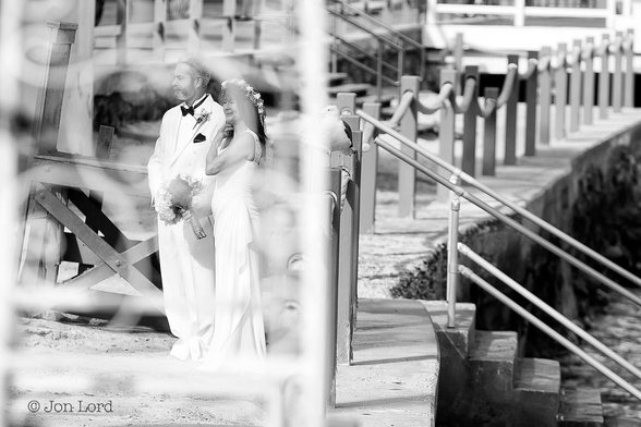 This is a black and white street photo in landscape format of a couple standing on the waterfront and about to be married. Avalon, Santa Catalina (2014).

In the foreground there is some white metal framework with piping delicately bent into circles, these are close to the camera and heavily blurred out of focus. Stretching away from the camera is a low, stone harbour wall that goes from the centre of the frame and arches around to the right. Along the wall are wooden posts with heavy hemp rope acting as a safety guard. There is a set of concrete steps that descend from the centre of the image down to the right and into the sea. Standing to the left of centre and in the middle ground is a middle aged couple, both viewed partially in profile, the lady closest to the camera and the man, to her right. The lady is wearing a long white dress and the man a white dinner jacket suit (tuxedo) with a black bow tie. The lady has a ring of flowers on the top of her hair and a bunch of roses in her hands. 
The metal work in the foreground almost forms a heart shape over the couple.
They are both waiting for the marriage ceremony to start.