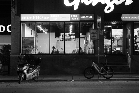Night. Across the street, the window of Ginger restaurant is brightly lit, showing the customers at the tables inside. Signs on the facade offer bubble tea, smoothies, the best Pho ever. On the street outside there’s a parkedmoped at left and bicycle locked to a post at right. Both are mostly in the dark, lit only by the street lights and traffic.