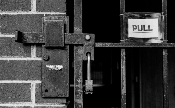 A black and white photo of a gate latch on a red brick wall with a padlock securing it.  On the bars of the gate there's a zip tied sign that reads "PULL".