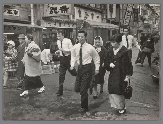 The image depicts a bustling street scene in Japan from the early to mid-1960s, as indicated by the clothing style and architecture. A group of people is captured in motion on what appears to be a busy pedestrian road lined with shops displaying Japanese characters.

In the foreground, there are two women walking side-by-side; one dressed in traditional attire including a kimono and geta sandals, while her companion wears Western-style clothes. Behind them strides an older man wearing glasses, followed by three individuals: a couple holding hands — the woman's head is adorned with a white veil or bonnet, suggesting she may be married.

The men ahead are both dressed in business attire; one prominently features dark hair and glasses. The scene is filled with activity as other pedestrians move about their daily routines. Some carry items like baskets, while others walk alone carrying bags over the shoulder.

The backdrop showcases an urban environment with various storefronts displaying Japanese text on signs. Notable details include a sign for "Kanji" in bold letters above one of the shops and another that reads "Edo." The architecture suggests mixed-use buildings typical of commercial areas, likely containing both businesses and residences.

Overall, this photograph by Ed van der Elsken captures an authentic slice of life from Japan during its period of significant urban development.