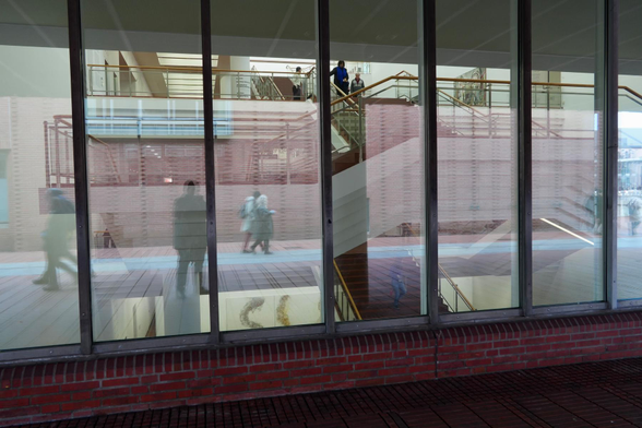 People are walking on stairs inside a building with reflections visible on large glass windows from the outside.