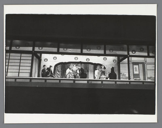 The image captures a dramatic scene from what appears to be a traditional Japanese theater performance, specifically in the Bunraku style. This form of puppetry is renowned for its intricate masks and puppets that represent various characters on stage.

In this composition, we see multiple figures positioned behind barriers with decorative motifs resembling chrysanthemum flowers and circular patterns, which could symbolize status or rank within the performance context. These performers are dressed in elaborate costumes indicative of historical or mythological roles; some wear robes adorned with ornate designs, suggesting nobility or high-ranking characters.

The puppets have a lifelike quality due to their detailed clothing and accessories, such as headpieces that signify different social statuses. The setting includes architectural elements like sliding doors (shoji) common in traditional Japanese architecture, enhancing the authenticity of the scene depicted.

This black-and-white photograph accentuates contrasts between light and shadow, adding depth and texture to both the performers' costumes and their surroundings. Overall, it offers a glimpse into Japan's rich theatrical heritage and its meticulous attention to detail in storytelling through puppetry.