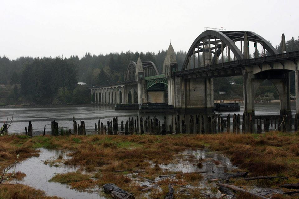 On a moody, overcast day a Deco bridge is seen over a river. On the near bank pf the river are well-weathered old posts and soggy wetland grasses.