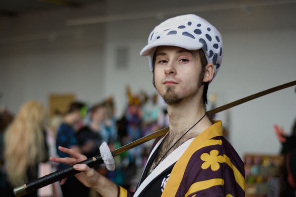 The image captures a young man who appears to be cosplaying as Trafalgar Law from the anime "One Piece". He is dressed in an yellow and purple kimono with yellow flowers. On his head, he sports a distinctive hat adorned with black polka dots.

In his right hand, he holds a sword, its blade is placed on his shoulder.

The background suggests an indoor setting with other people present, possibly indicating that this photo was taken at a convention or similar event where such activities are common. However, these individuals and their details are blurred, keeping the focus on our cosplay subject.