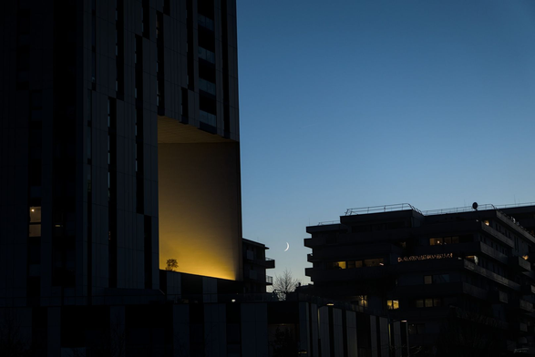 Das Foto zeigt eine städtische Szene während der Dämmerung oder des frühen Abends. Im Vordergrund sind moderne Gebäude mit dunklen Silhouetten zu sehen, die sich gegen den blauen Himmel abheben. Besonders auffällig ist ein schmales, hohes Gebäude links im Bild, das eine Einbuchtung hat und von einer Lichtquelle angestrahlt wird und dadurch eine goldene Fassade aufweist. Zwischen den Gebäuden ist der Mond als schmale Sichel sichtbar. Die Atmosphäre wirkt ruhig und fast mystisch durch das sanfte Licht und die klaren Linien der Architektur.