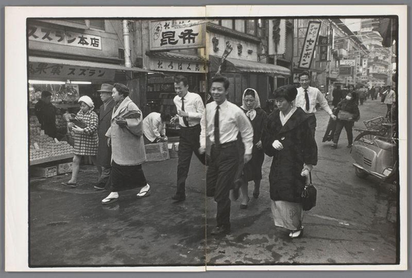 The image captures a bustling street scene in an urban setting. Several individuals of varying ages and genders are captured mid-stride, suggesting movement through the space. The people appear to be engaged in daily activities such as walking or shopping, indicative of everyday life within this locale.

In terms of attire, some wear traditional clothing like kimonos, hinting at a cultural context where both modernity and tradition coexist. Others are dressed more formally with ties and coats, which could suggest they're either on their way to work or returning from it.

The architecture in the background includes storefronts with signage that appears non-Latin script-based, typical of Asian street settings like those found in Japan. The presence of such stores along with other elements suggests a commercial area within an urban environment.

Overall, this image reflects daily life and culture as experienced on a specific city street during what could be early to mid-20th century Japanese era judging by the fashion styles and black-and-white photography technique used here.
