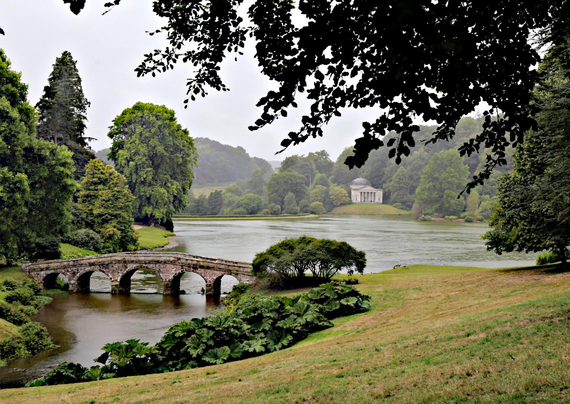 A very famous scene at Stourhead,  photographed from under a tree near the entrance to the walk around the lake.
Front left, a bridge, with the central lake extending though 5 visible arches to the narrower inflow in the bottom left corner.
In the distance a temple modelled on the Pantheon in Rome, the lake before it, and trees and then sky beyond it.  There's a grassy slope in the right foreground, and rain is visible on the water surface.
