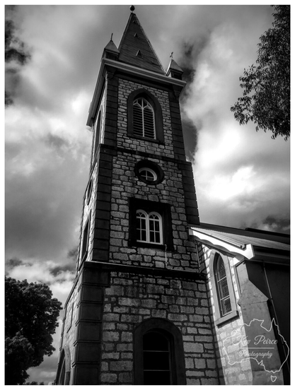 Dramatic, low angle black and white photograph focusing on the tall, stone bell tower and spire of the Kapunda Catholic Church in South Australia.  The tower features dark, textured stonework and arched windows, contrasting sharply against a heavily clouded and moody sky.