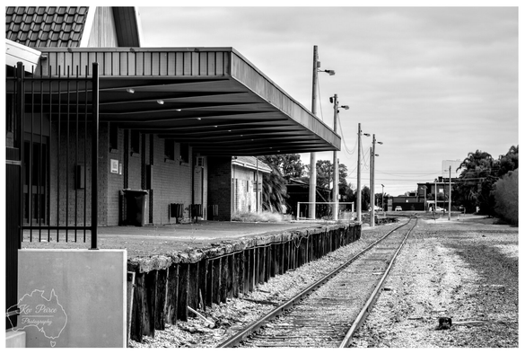 Black and white photograph showing a long view down the unused tracks and platform of Mildura Railway Station.

The platform, made of wood and brick, extends from the left under a cantilevered metal awning. The rails recede sharply into the distance under an overcast sky. The image conveys a sense of stillness and historical solitude.