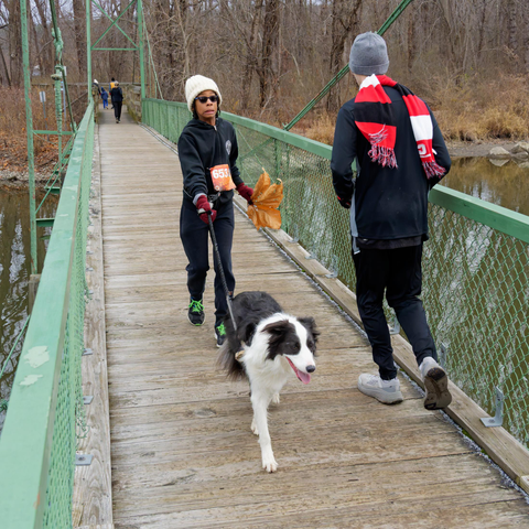 Women in black jacket and a white wool hat  runs with a black and white dog across a suspension bridge with another runner going the other way.
