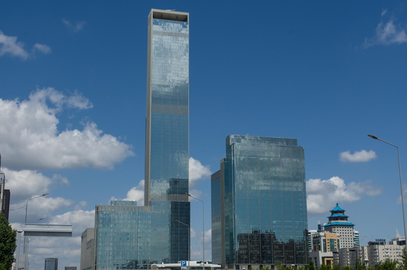 The image depicts a cityscape with a tall, modern glass skyscraper dominating the skyline. The building's reflective surface mirrors the surrounding environment and sky, creating an interesting visual effect. In front of the skyscraper, there is another building that appears to be older in design but still stands out due to its unique architectural features.

The scene also includes several other buildings scattered throughout the cityscape, showcasing a mix of old and new structures. The sky above is clear blue with fluffy white clouds, adding depth and dimension to the image.
