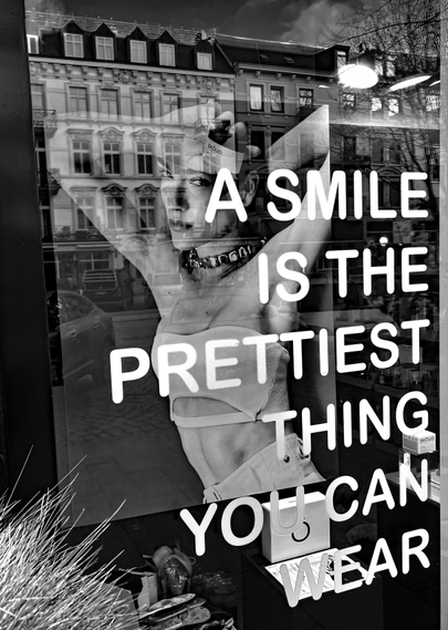 Ein Schaufenster mit der aufgeklebten Botschaft: "A smile is the prettiest thing you can wear", dahinter ein Werbeplakat mit einer jungen Frau, sie schaut eher grimmig-distanziert, unfreundlich. Exakt das Gegenteil zur Botschaft. In der Fensterscheibe spiegeln sich Jugendstilhäuser der gegenüberliegenden Straßenseite mit vielen Fenstern. Das Bild ist schwarz-weiß.

A shop window with a sticker that reads: ‘A smile is the prettiest thing you can wear.’ Behind it is an advertising poster featuring a young woman who looks rather grim and distant, unfriendly. Exactly the opposite of the message. The window pane reflects the Art Nouveau buildings across the street with their many windows. The image is black and white.