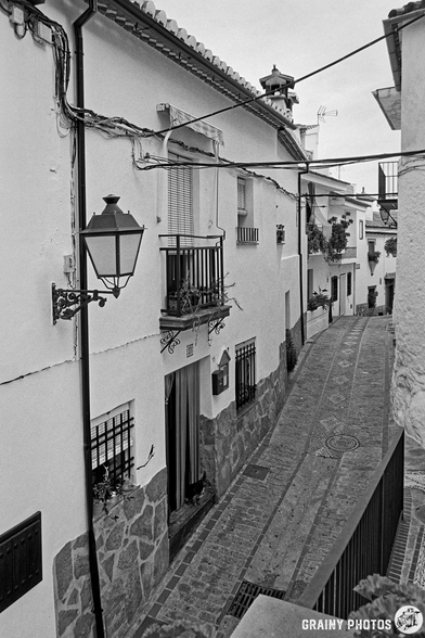 A narrow, cobblestone alleyway lined with white houses, featuring decorative balconies and hanging plants. Vintage street lamps illuminate the space, creating a charming atmosphere under a cloudy sky.
