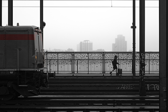 Bild von einer Eisenbahnbrücke, im Vordergrund Teil einer Lok, dahinter eine Person mit Rollkoffer, im Hintergrund-Nebel Hochhäuser am Horizont