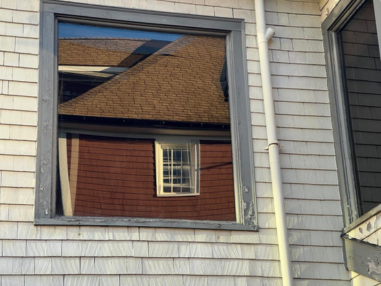 A window reflects several images of a wooden building, and the images are slightly offset and distorted relative to one another. The building in the image has maroon wooden siding with white trim around alameda window, and a sloping brown shingled roof.  The window itself is framed by peeling grey trim ona white clapboard building. The right side of the image also shows a white drainpipe and another window facing a different direction, with no reflection. [Halifax (NS, Canada) November 2025]