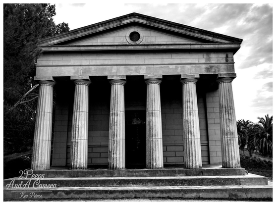 A low angle, black and white photograph of the imposing Seppelt Family Mausoleum at Seppeltsfield in the Barossa Valley. 

The Grecian style structure features six large Doric columns across the front, supporting a triangular pediment with a central round window (oculus).

Stone steps lead up to the portico. The dark shadows contrast sharply with the white stone and bright, cloudy sky.