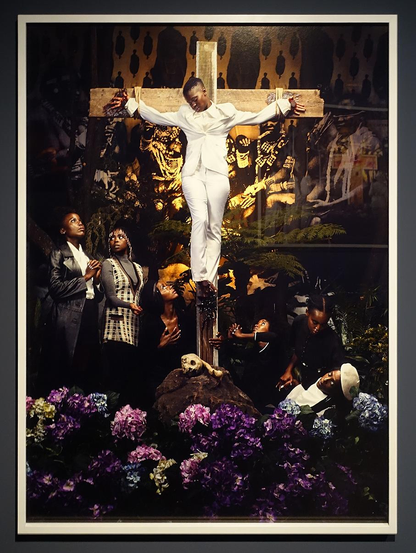 Een donkere man in een wit pak hangt aan een houten kruis dat in de schijnwerpers staat. In het donker daarachter een collega afbeeldingen van voorouders. Links naast het kruis staat een jong stel in devotie te kijken naar de man. Rechts verzorgt een vrouw een liggende man. Daarvoor een zee aan prachtig blauwpaarse bloemen.