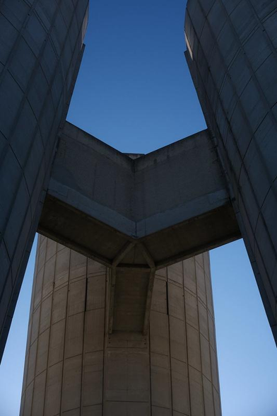 Photo en contre plongée d’une structure en béton à 3 branches reliant 3 autres gros bâtiment en béton aussi dont on ne voit qu’une petite partie, sur fond de ciel bleu