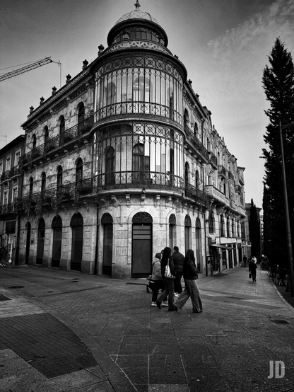 Una fotografía en blanco y negro de una calle urbana. En el centro, un gran edificio de piedra ornamentado se alza en la esquina de una calle adoquinada. Su característica más distintiva es una sección curva y acristalada en la esquina, que se eleva varios pisos y termina en una pequeña cúpula. El edificio tiene múltiples ventanas con balcones de hierro forjado y arcos en la planta baja. En primer plano, un grupo de personas camina por la acera hacia la esquina del edificio. A la derecha, árboles altos y oscuros se alinean en la calle. En el horizonte de la izquierda, se vislumbra el brazo de una grúa. La luz del día crea fuertes contrastes de luces y sombras en la arquitectura y el pavimento.