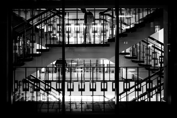The image is a black and white photograph showcasing an architectural interior, likely a building's entrance or lobby, viewed through a large window. The central focus is a set of multi-level staircases, which create a striking geometric pattern. The stairs are dark, likely made of wood, and they ascend and descend in a crisscrossing pattern. The railings are also dark, with a series of rectangular cutouts that add to the visual complexity.

In the mid-ground, a person is standing on a landing, silhouetted against a bright background that suggests an illuminated interior space. The person's form is indistinct, adding a sense of mystery.